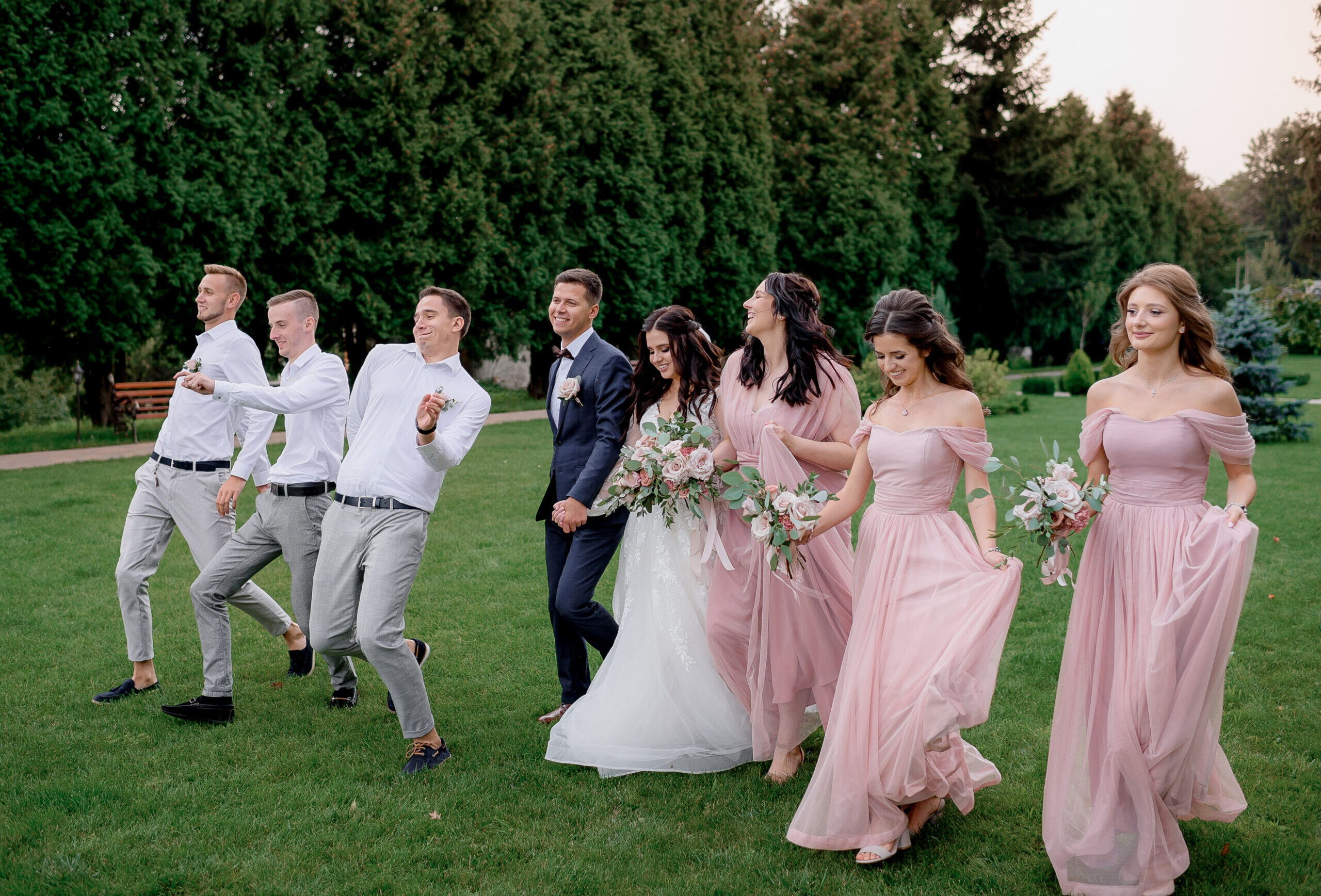 Bridesmaids dressed in pink dresses, best men and wedding couple are happily walking on the green yard